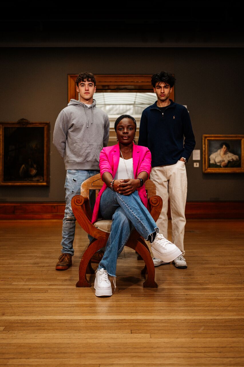 Three of Hull’s students sit and stand confidently together in a dark gallery with Georgian paintings in Ferens Art Gallery
