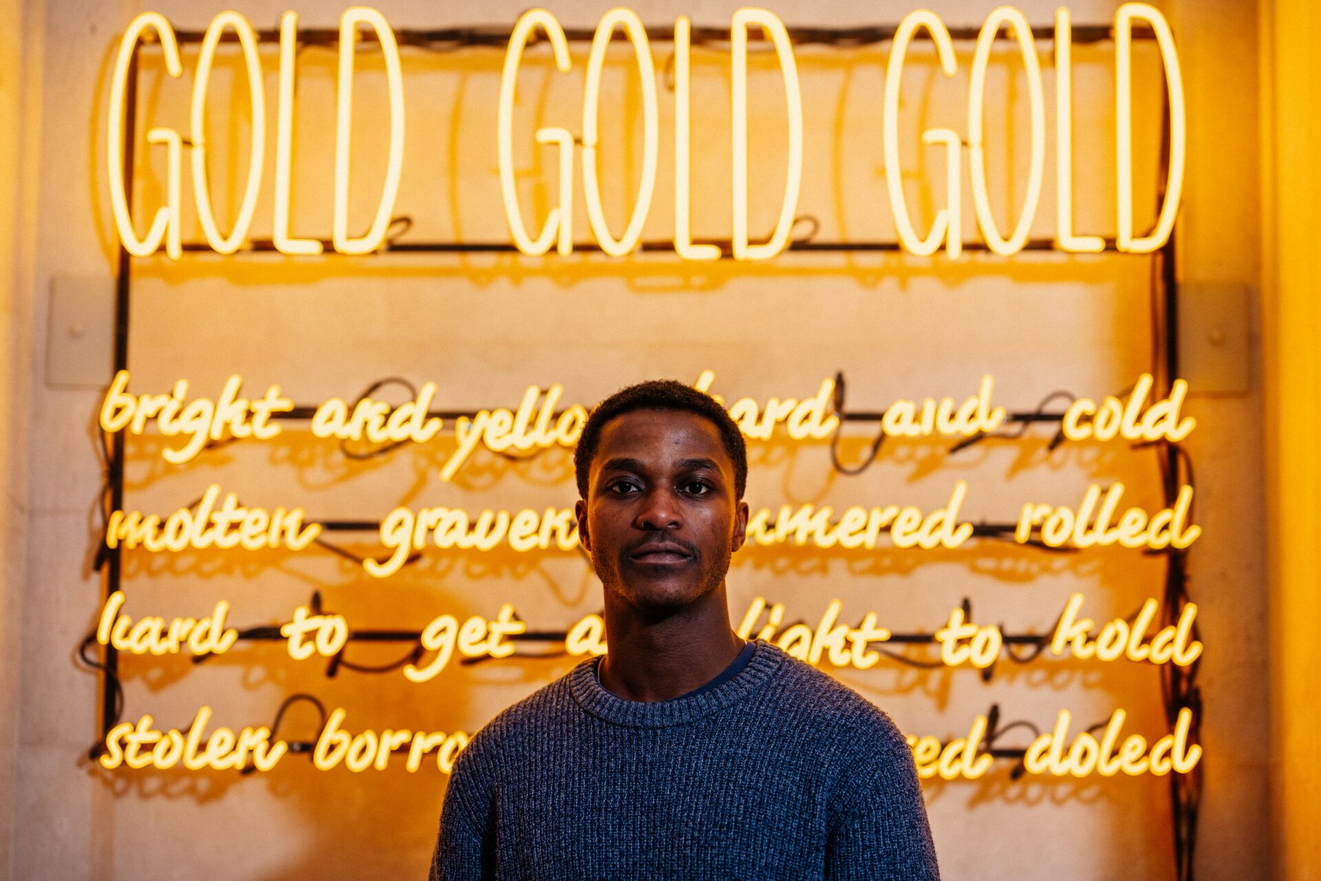 A male student in blue jumper stands in front of an artwork consisting of bright yellow tube lighting that says 'Gold Gold Gold'
