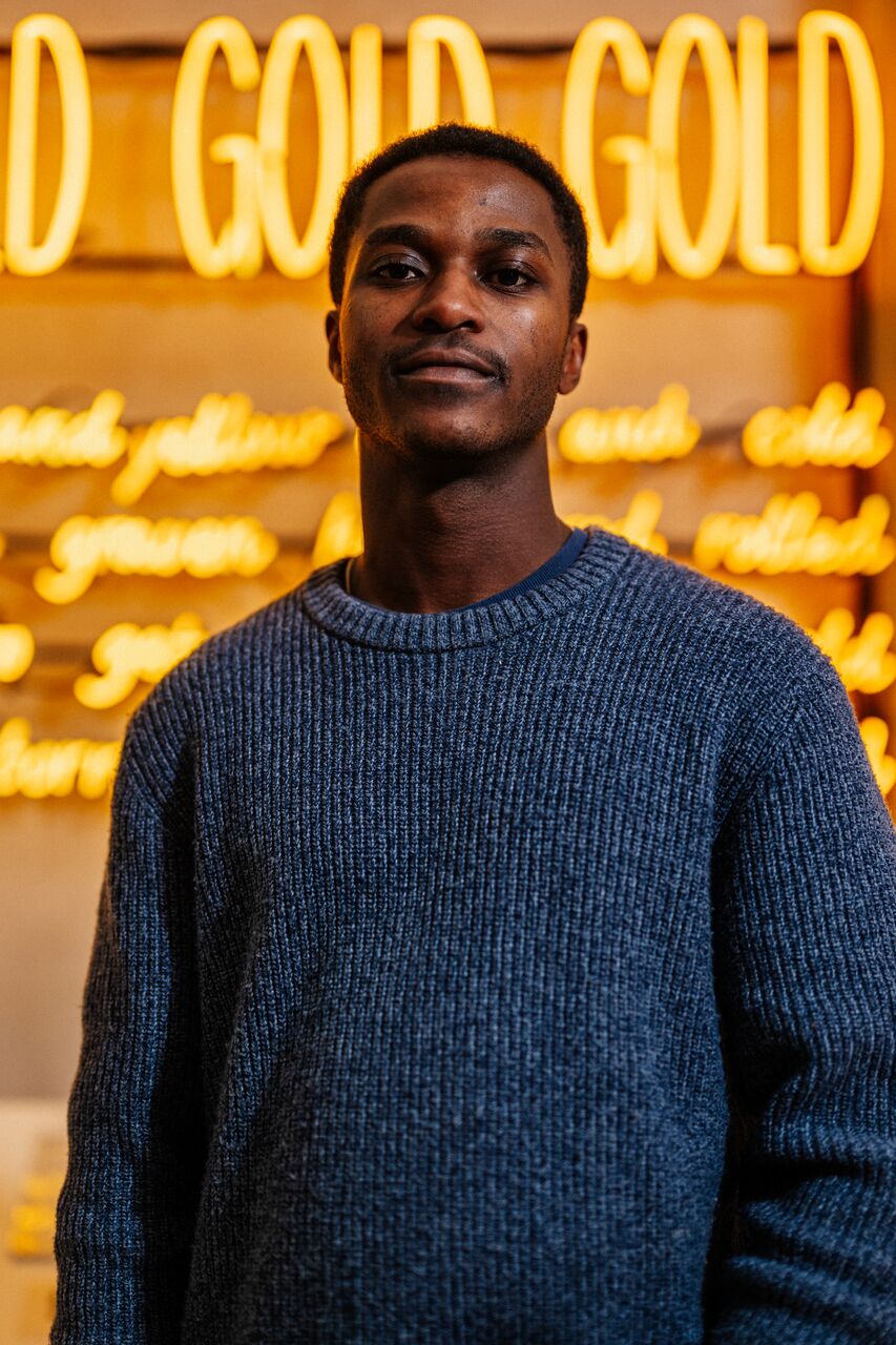 A male student in blue jumper stands in front of an artwork consisting of bright yellow tube lighting that says 'Gold Gold Gold'