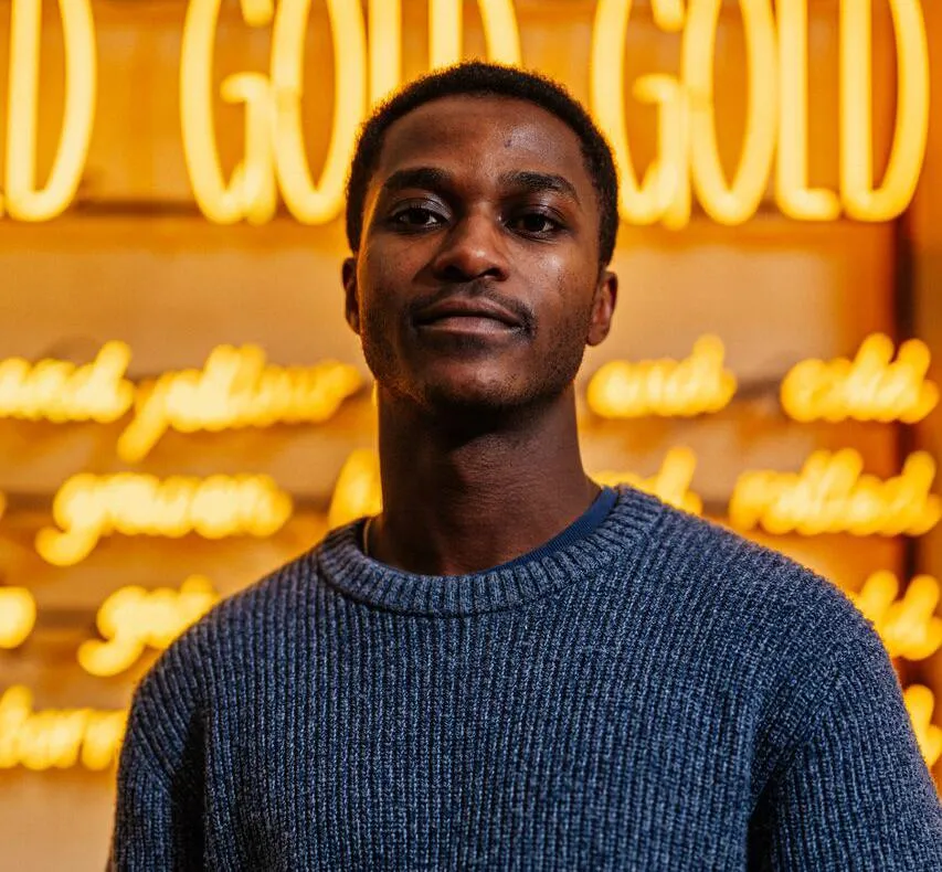 A male student in blue jumper stands in front of an artwork consisting of bright yellow tube lighting that says 'Gold Gold Gold'