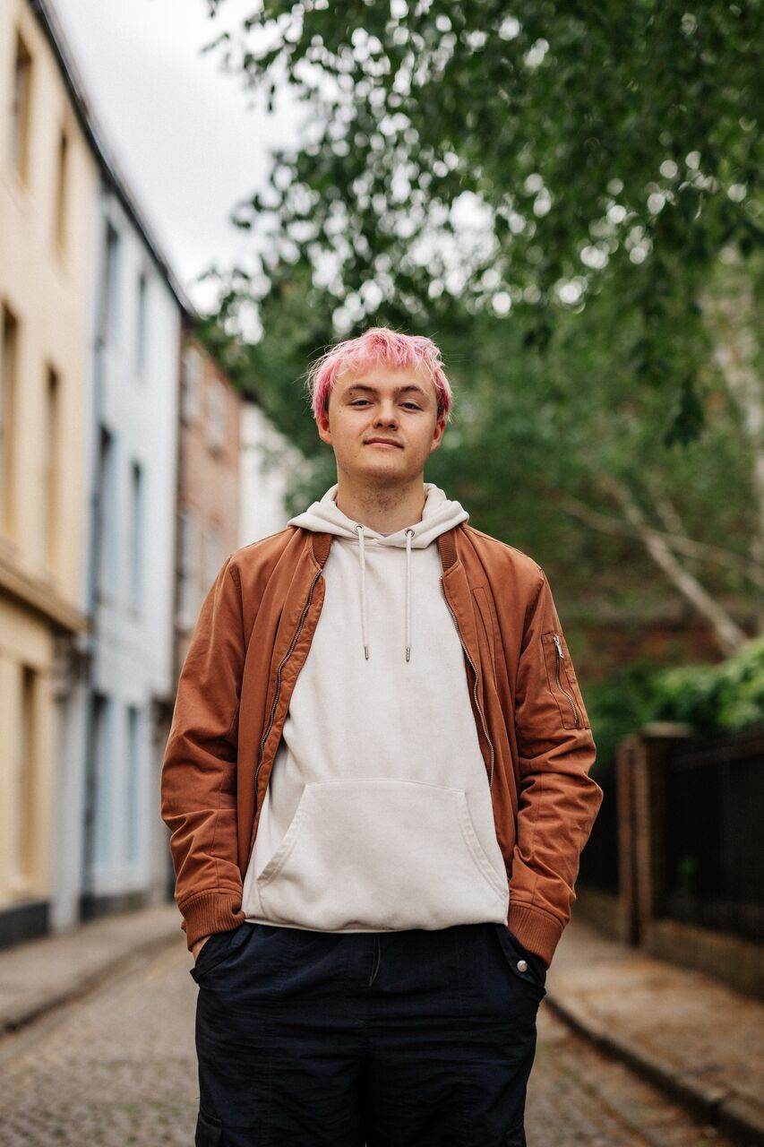 A male student with pink hair and brown jacket stands in a cobbled street with multicoloured houses