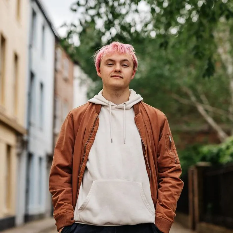 A male student with pink hair and brown jacket stands in a cobbled street with multicoloured houses