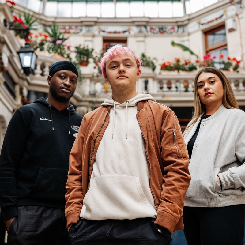 Three of Hull’s students stand confidently together in a beautiful shopping arcade with glass roof and flowers