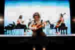 A music student tunes a violin while other musicians set up on a stage behind her