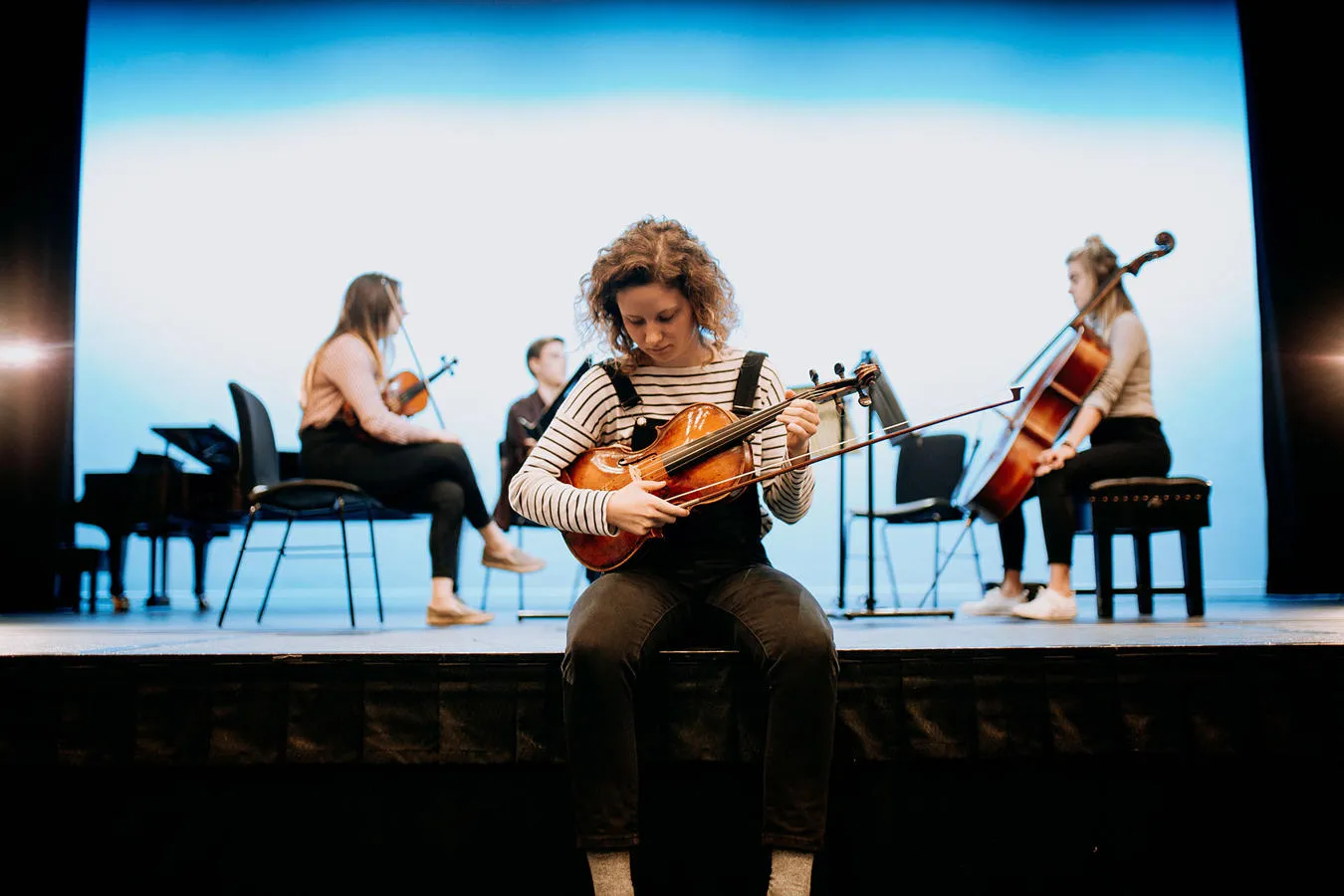 A music student tunes a violin while other musicians set up on a stage behind her