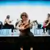 A music student tunes a violin while other musicians set up on a stage behind her