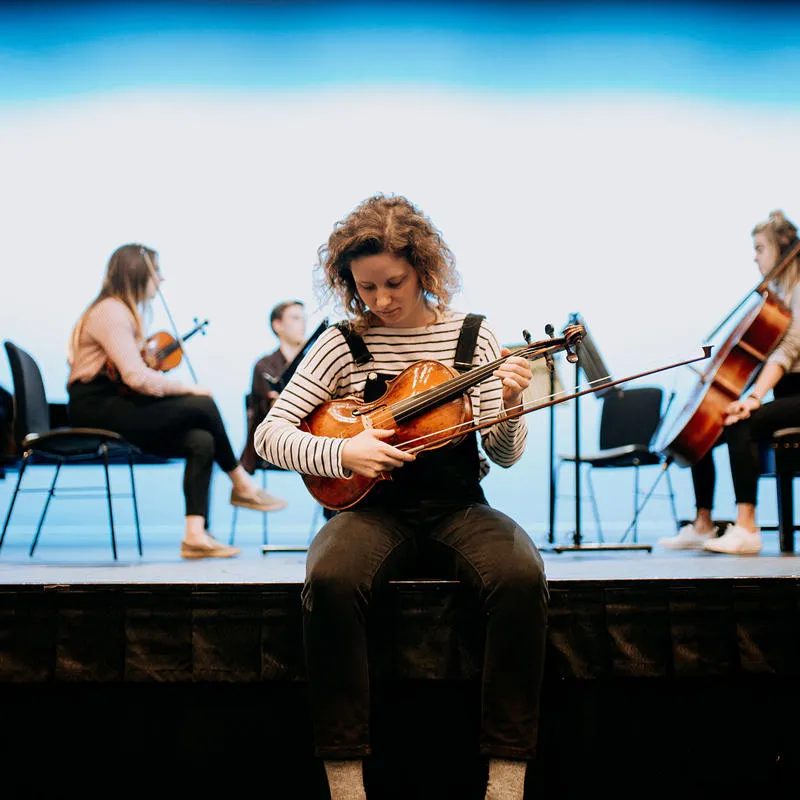 A music student tunes a violin while other musicians set up on a stage behind her
