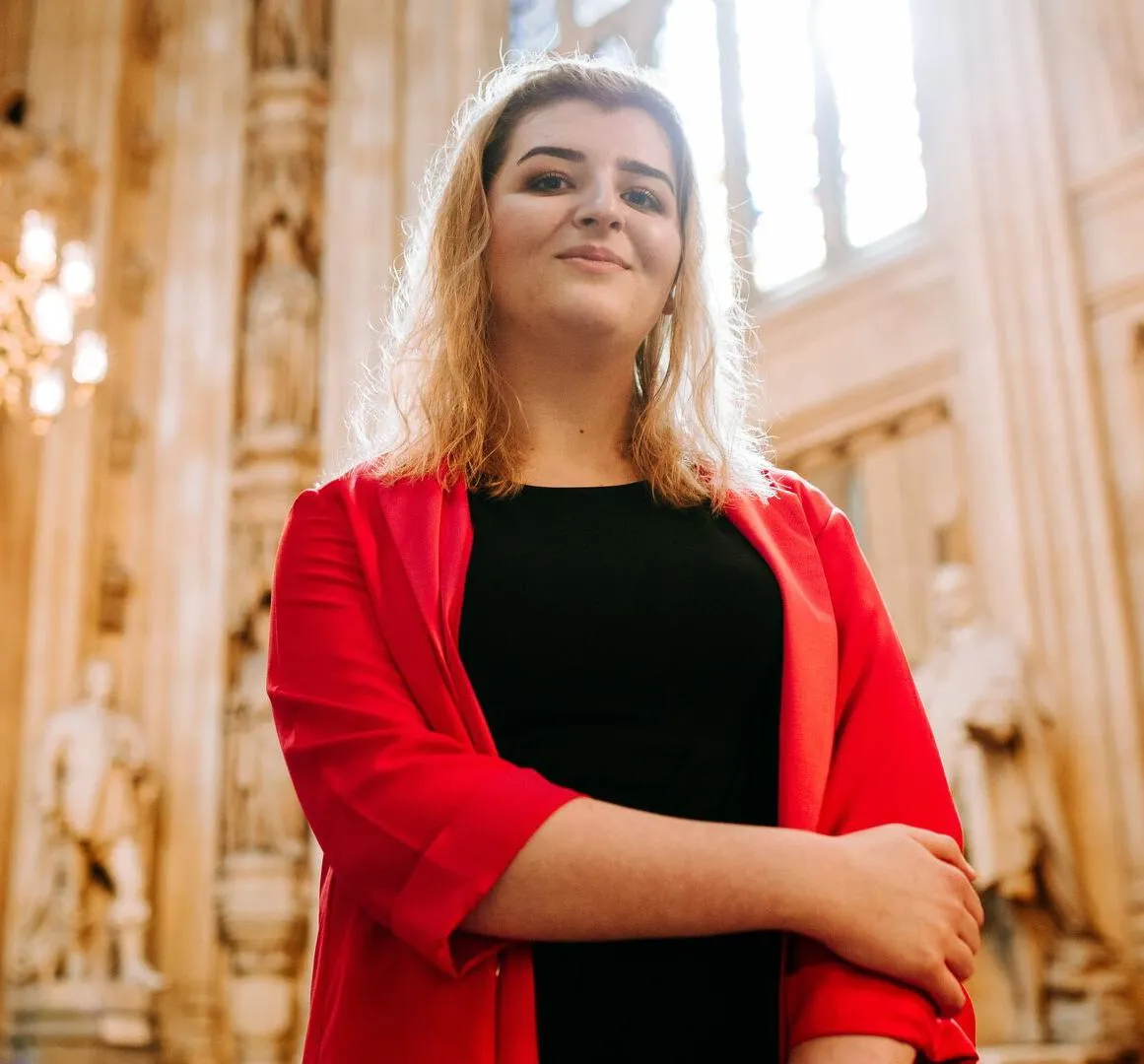 A female Politics student with blonde hair in a red blazer standing in the Houses of Parliament