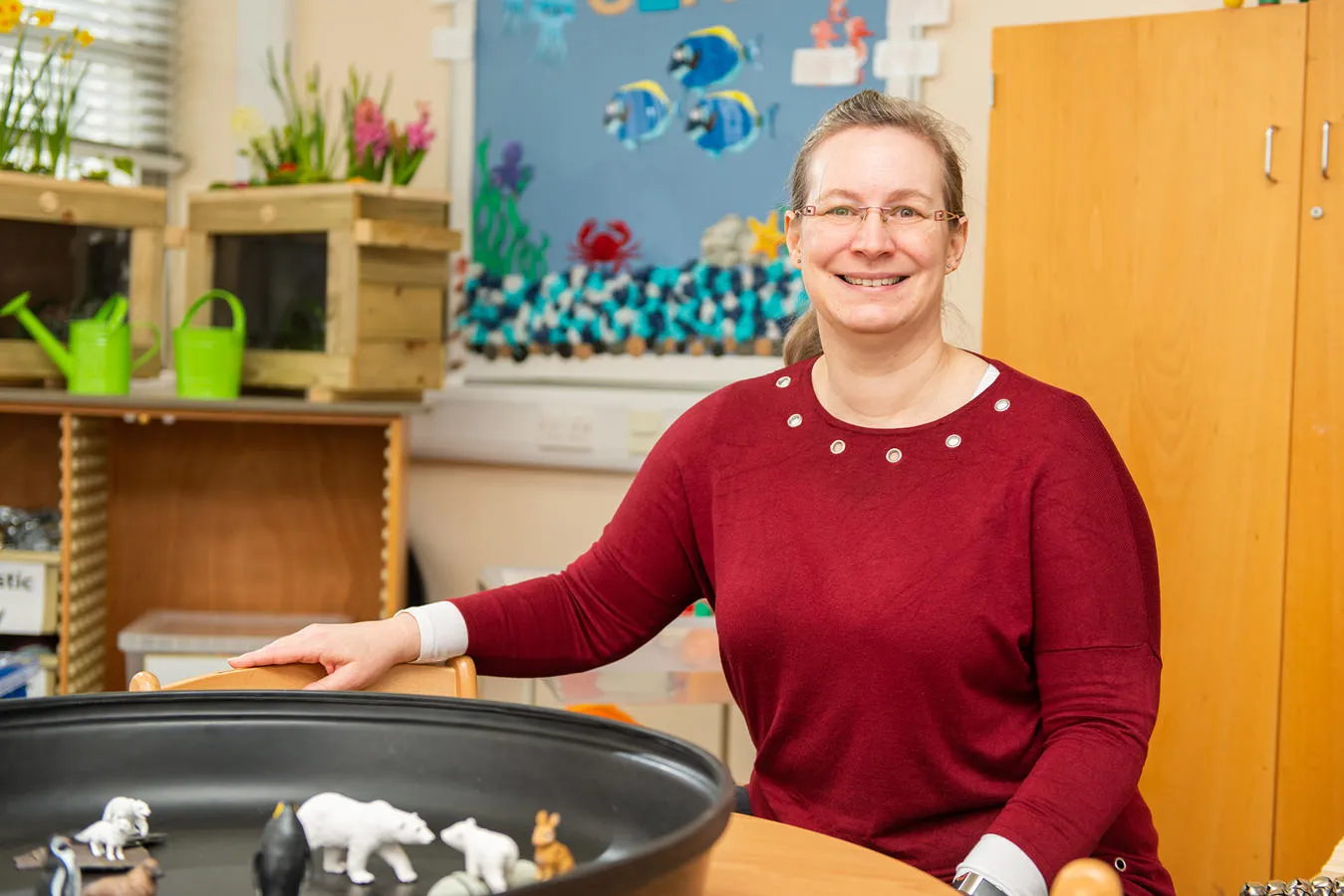 MA Education academic Cathy sits smiling in a teaching classroom with plastic animals on a table in front of her.