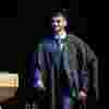 A male student strides confidently across the stage at Graduation in hat and gown.