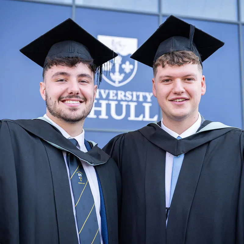 Two male graduates stand smiling in their gowns outside Connexin Live