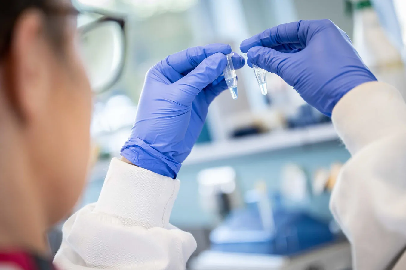 A woman in a white lab coat and blue gloves looks at two vials in a lab