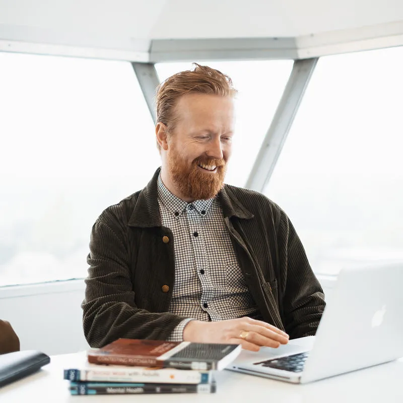 Masters student Chris works at a laptop on the seventh floor of the library, with a pile of books on the table in front of him.