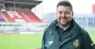 A paramedic wearing green NHS uniform standing by the side of a rugby pitch with a stand in the background