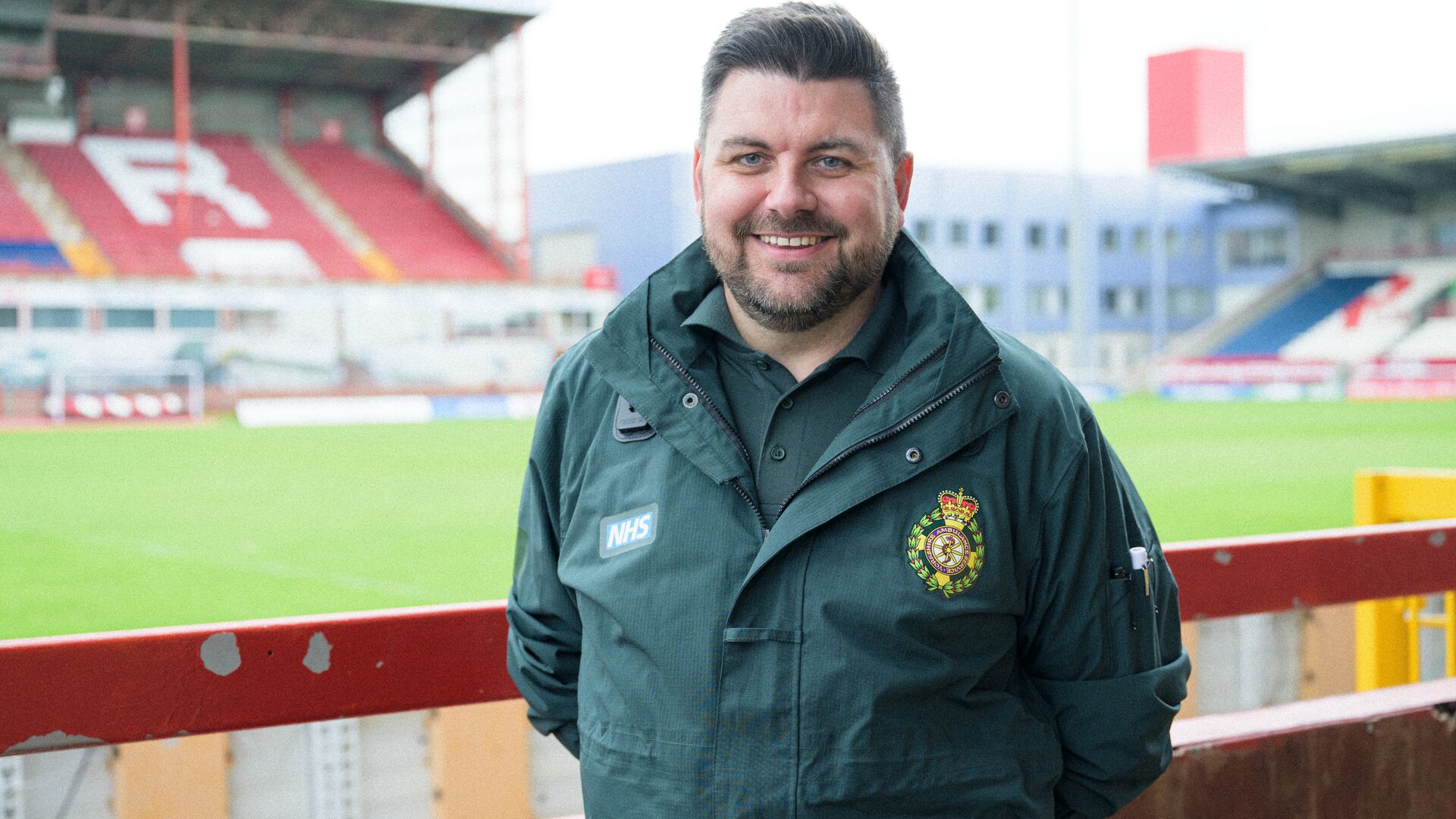 A paramedic wearing green NHS uniform standing by the side of a rugby pitch with a stand in the background