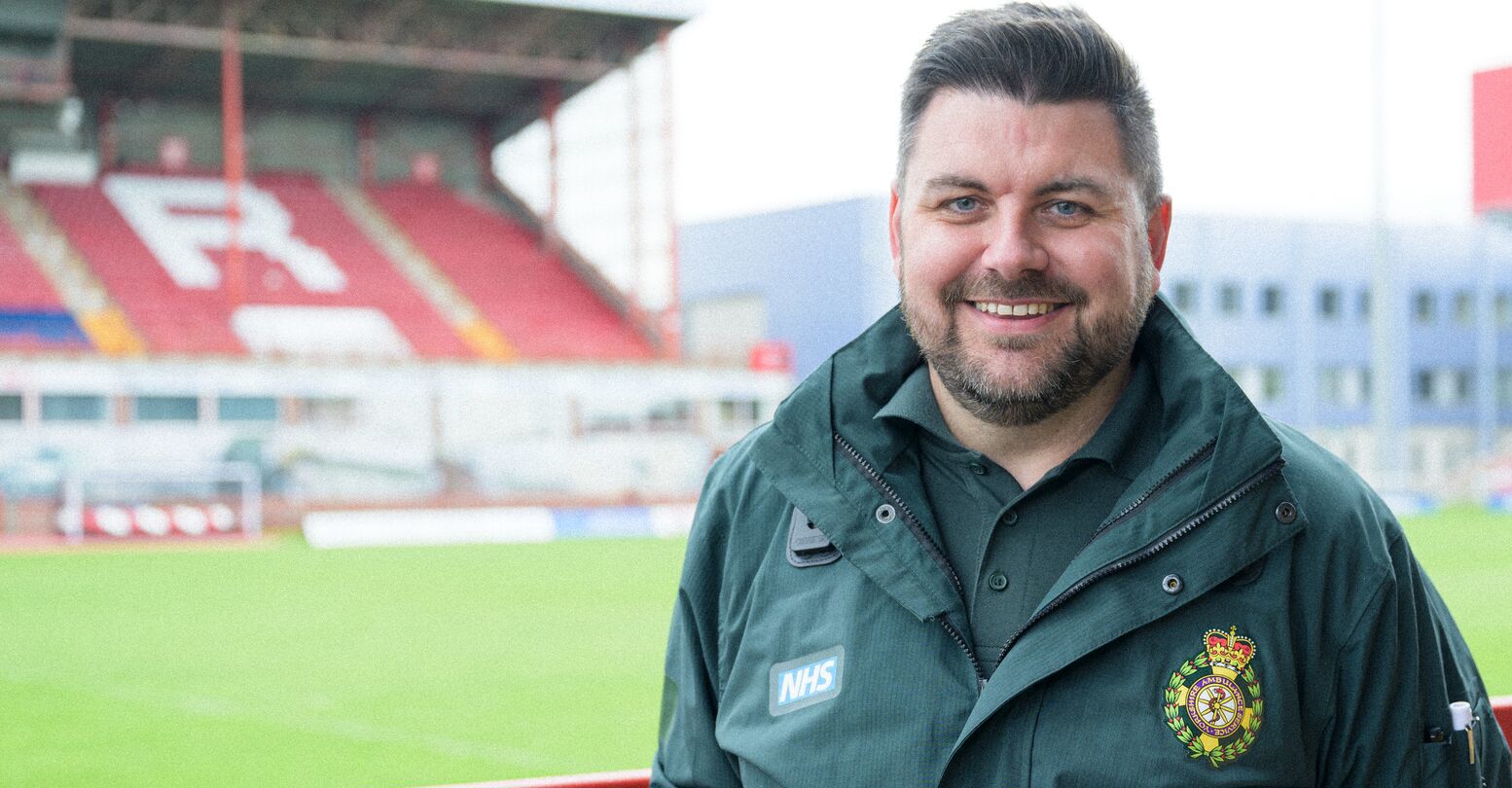 A paramedic wearing green NHS uniform standing by the side of a rugby pitch with a stand in the background