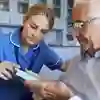 A community nurse wearing a blue uniform shows a patient a piece of equipment