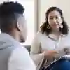 A woman sits with a pen and notepad listening to a young man in comfortable chairs