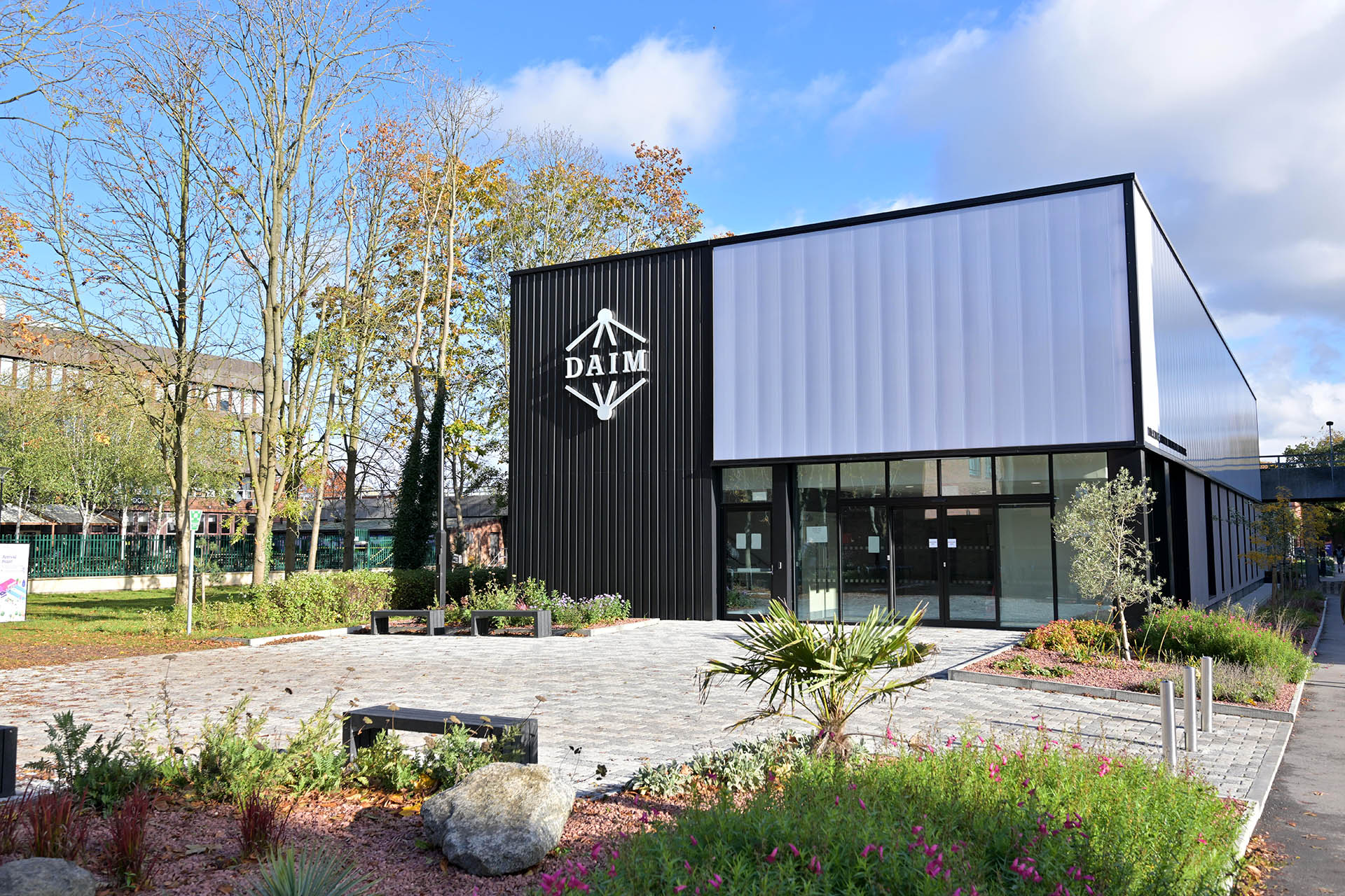 A modern black and white building with glass windows is surrounded by paving stones outlined by trees and bushes.