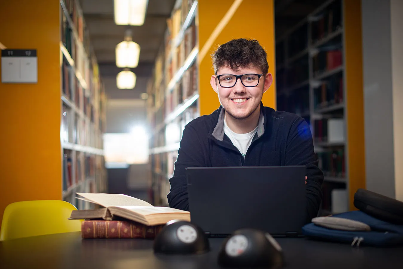 Student sits at a desk in a library with books and a laptop