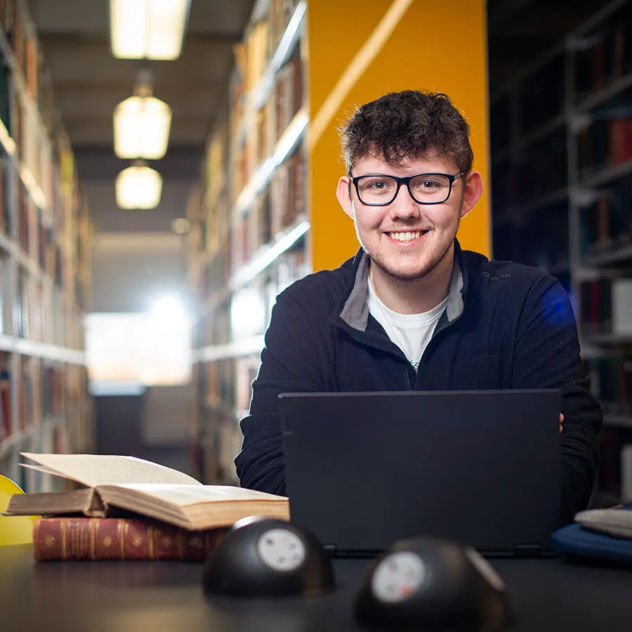 Student sits at a desk in a library with books and a laptop