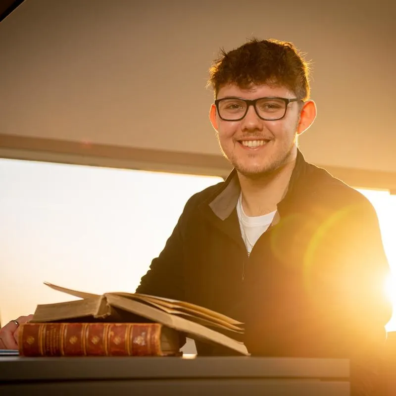 Hull History student, Dane Mellows, sits smiling with an open book and the sun resplendent behind him.