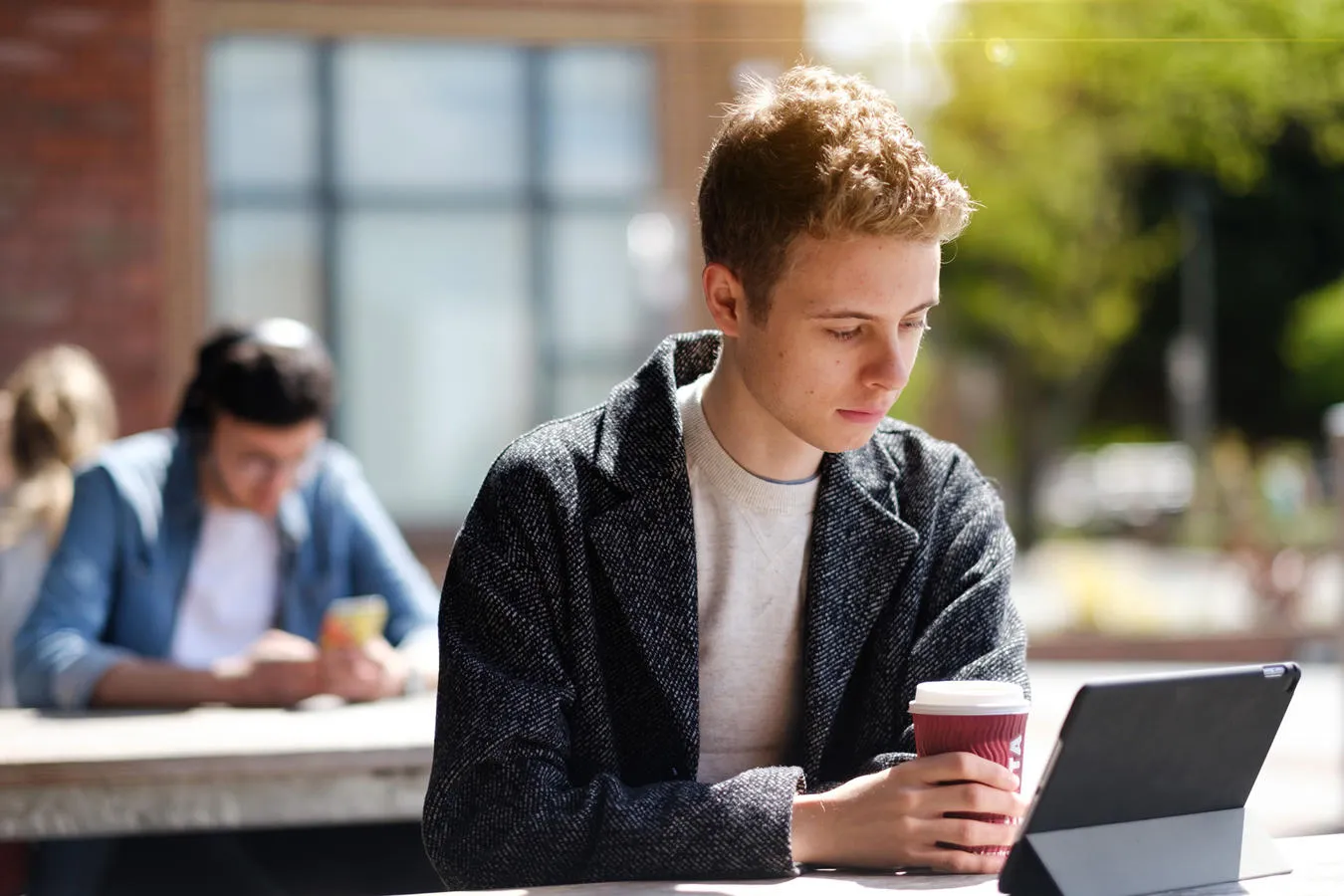 Student seated outdoors holding a coffee and looking at a tablet