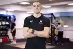Sports student Danny Uscroft-Mould stands, arms crossed, in a gym while other users workout on equipment behind him