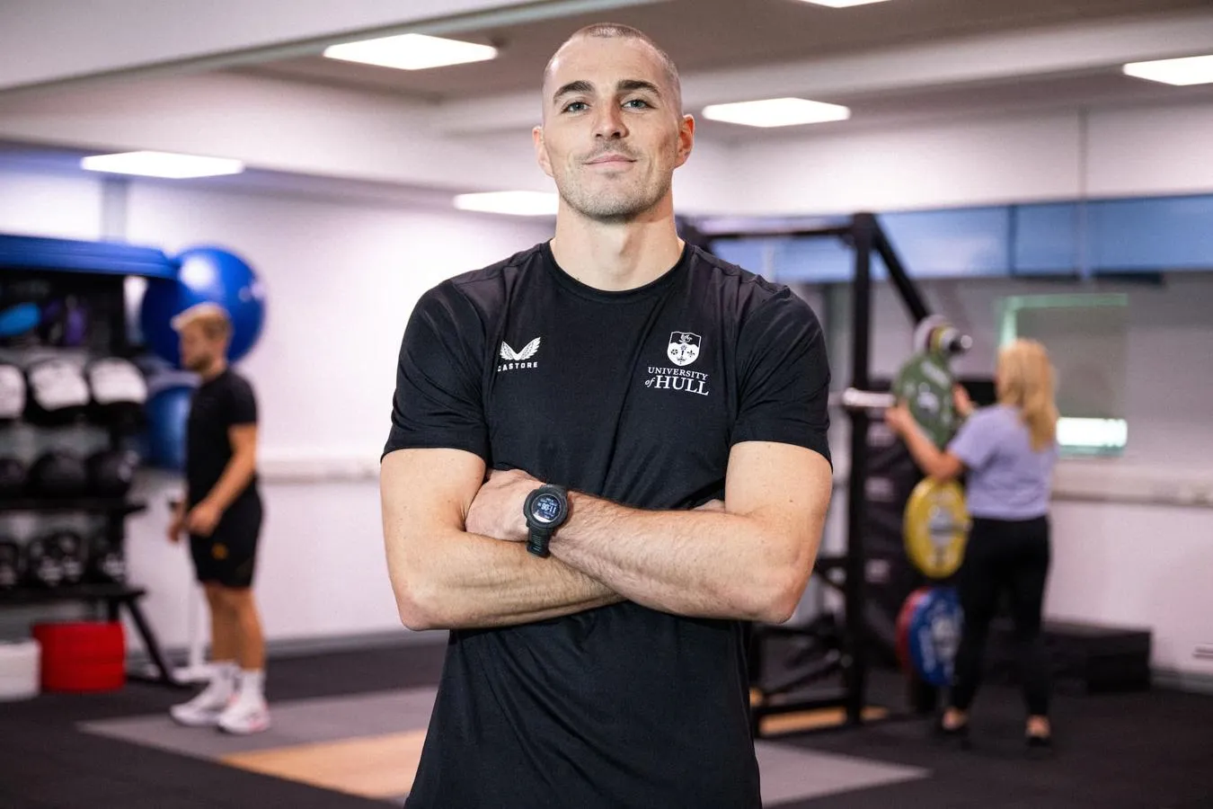 Sports student Danny Uscroft-Mould stands, arms crossed, in a gym while other users workout on equipment behind him