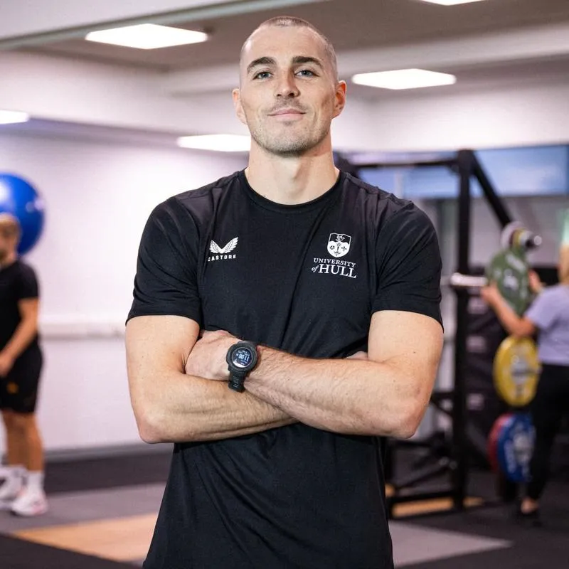 Sports student Danny Uscroft-Mould stands, arms crossed, in a gym while other users workout on equipment behind him