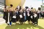A group of new graduates stand on the lawn outside Connexin Live waving their hats and holding degree certificates.