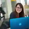 A young woman with glasses with a laptop sits in a room with large glass windows