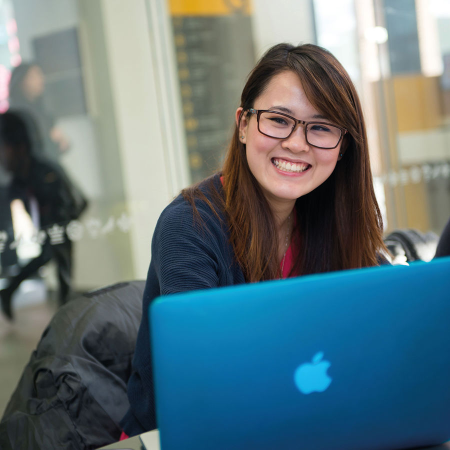 A young woman with glasses with a laptop sits in a room with large glass windows