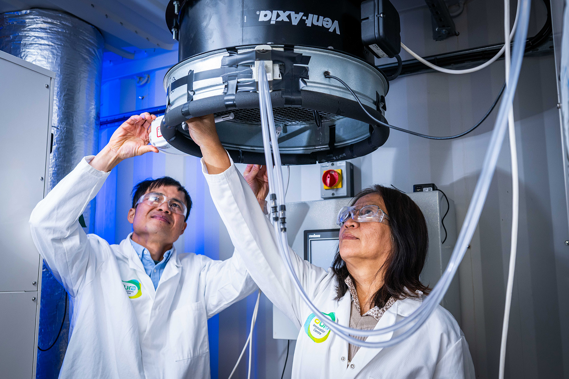 Two academics in white coats and safety glasses stand below a large circular vent in a lab space