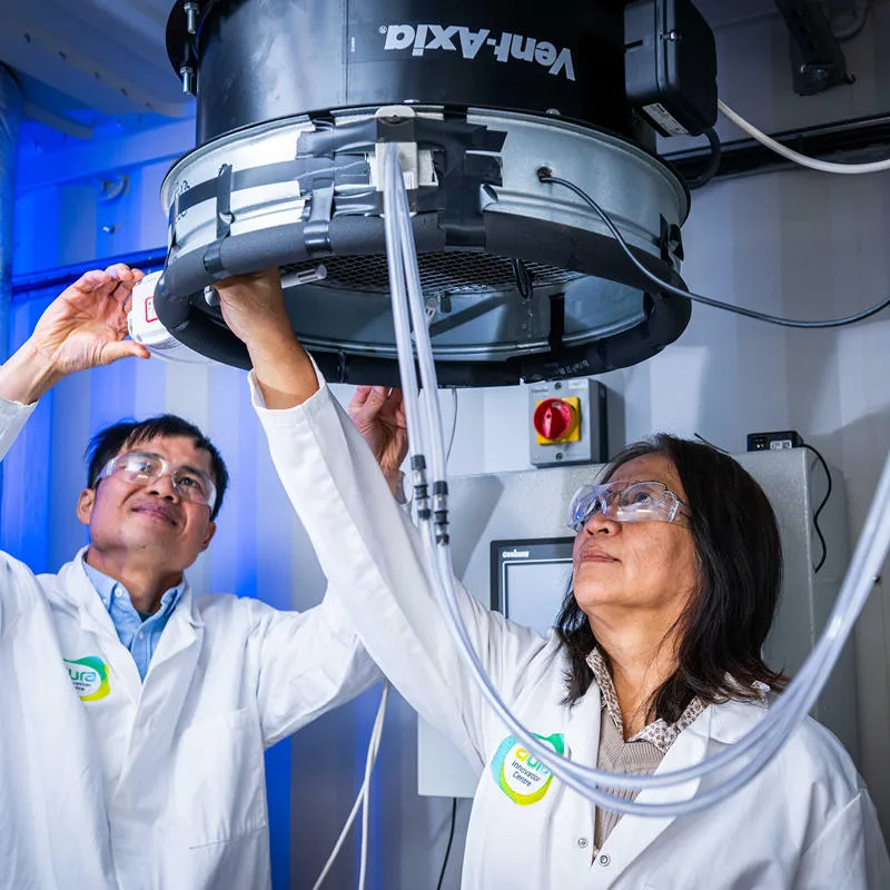 Two academics in white coats and safety glasses stand below a large circular vent in a lab space
