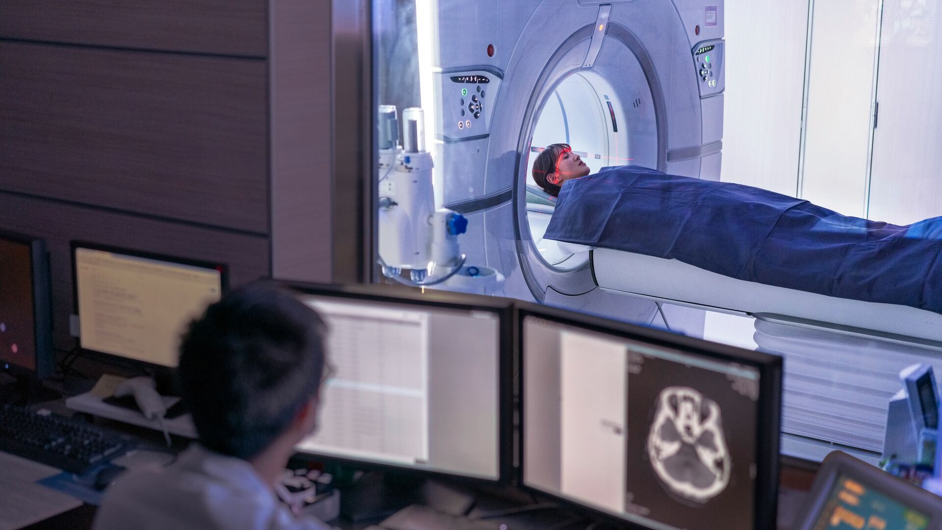 A doctor monitors a scan on a computer while in the background a patient enters an MRI scanning machine