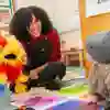 A teacher smiles at a baby while playing with a parrot puppet in a classroom