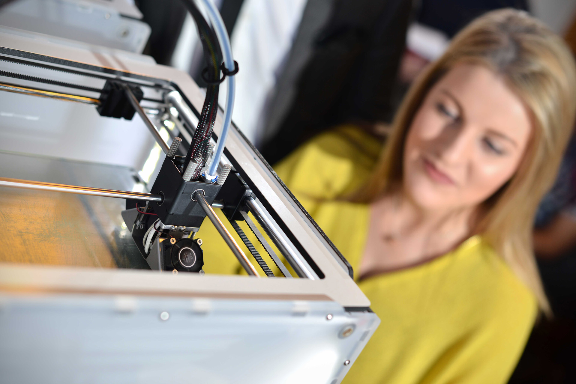 A 3D printer in the foreground with a female student looking on