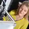 A 3D printer in the foreground with a female student looking on