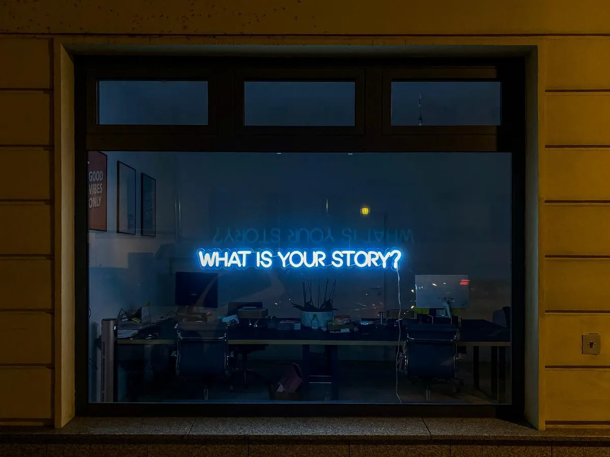 Neon sign in a dark window with the words 'what is your story?'