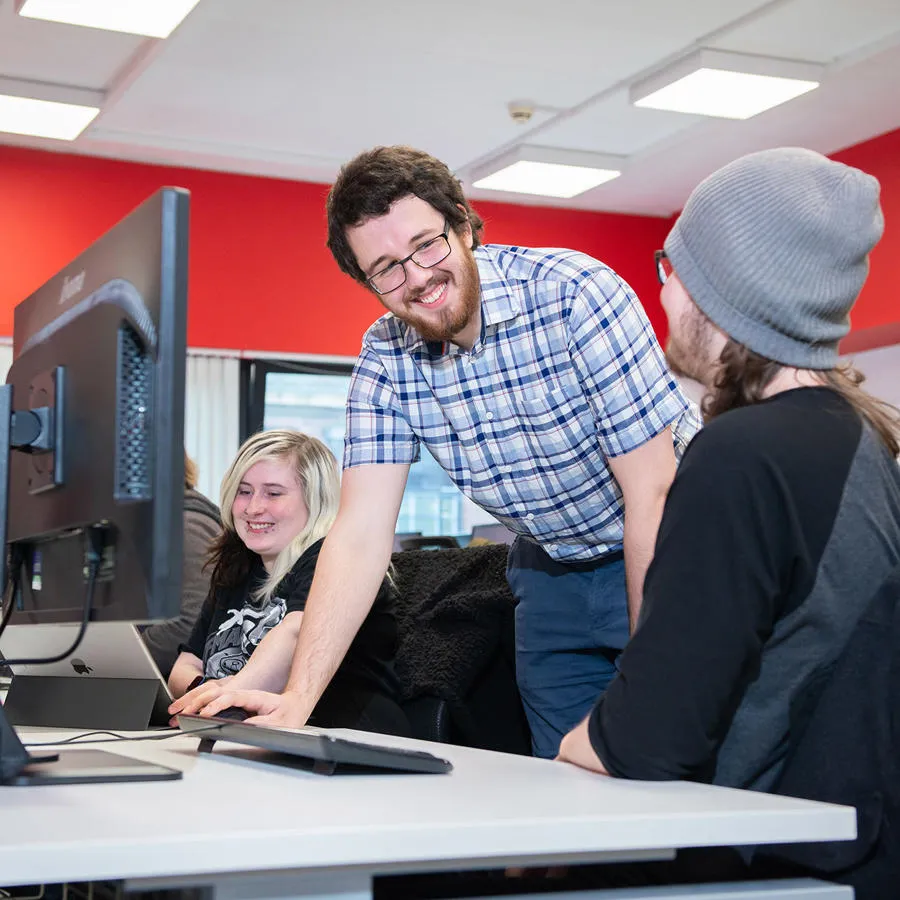 A male lecturer chats to two young adults in a computer studio