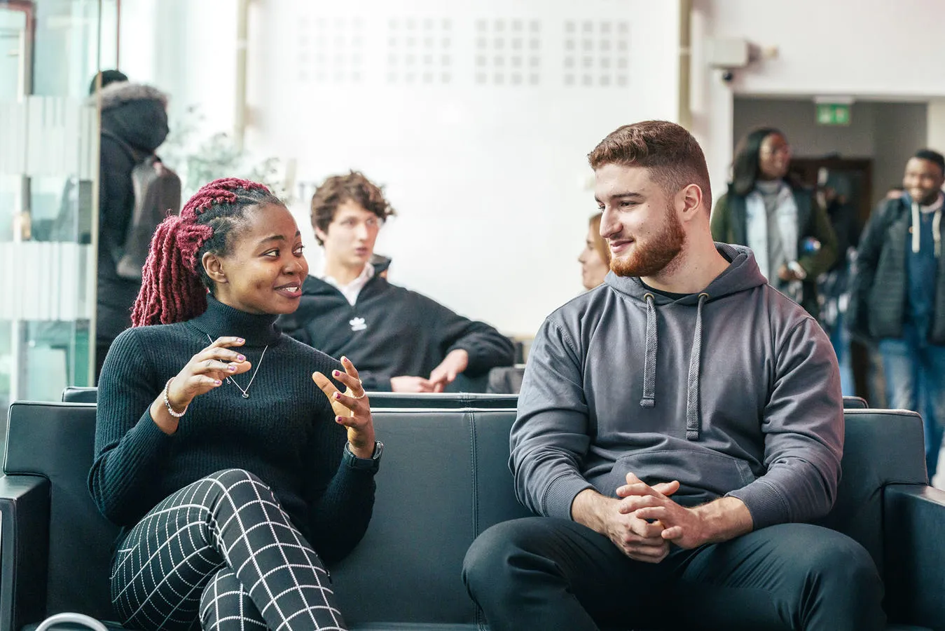 A female student and a male student sat on a black leather sofa chatting with people behind them engaging in their day to day activities.
