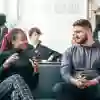 A young woman and man sit on a black leather sofa chatting with people behind them.