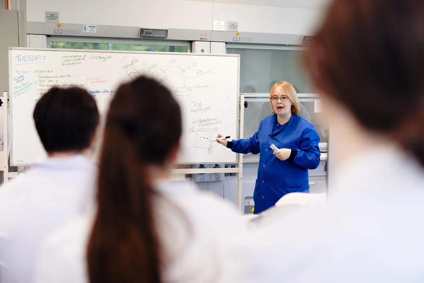 An instructor in a blue lab coat demonstrates on a whiteboard