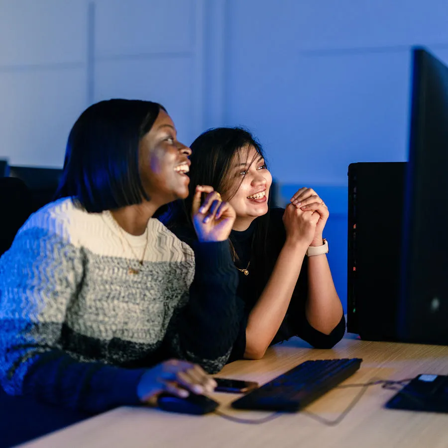 Two young woman chat and look at a computer screen in a dimly lit room