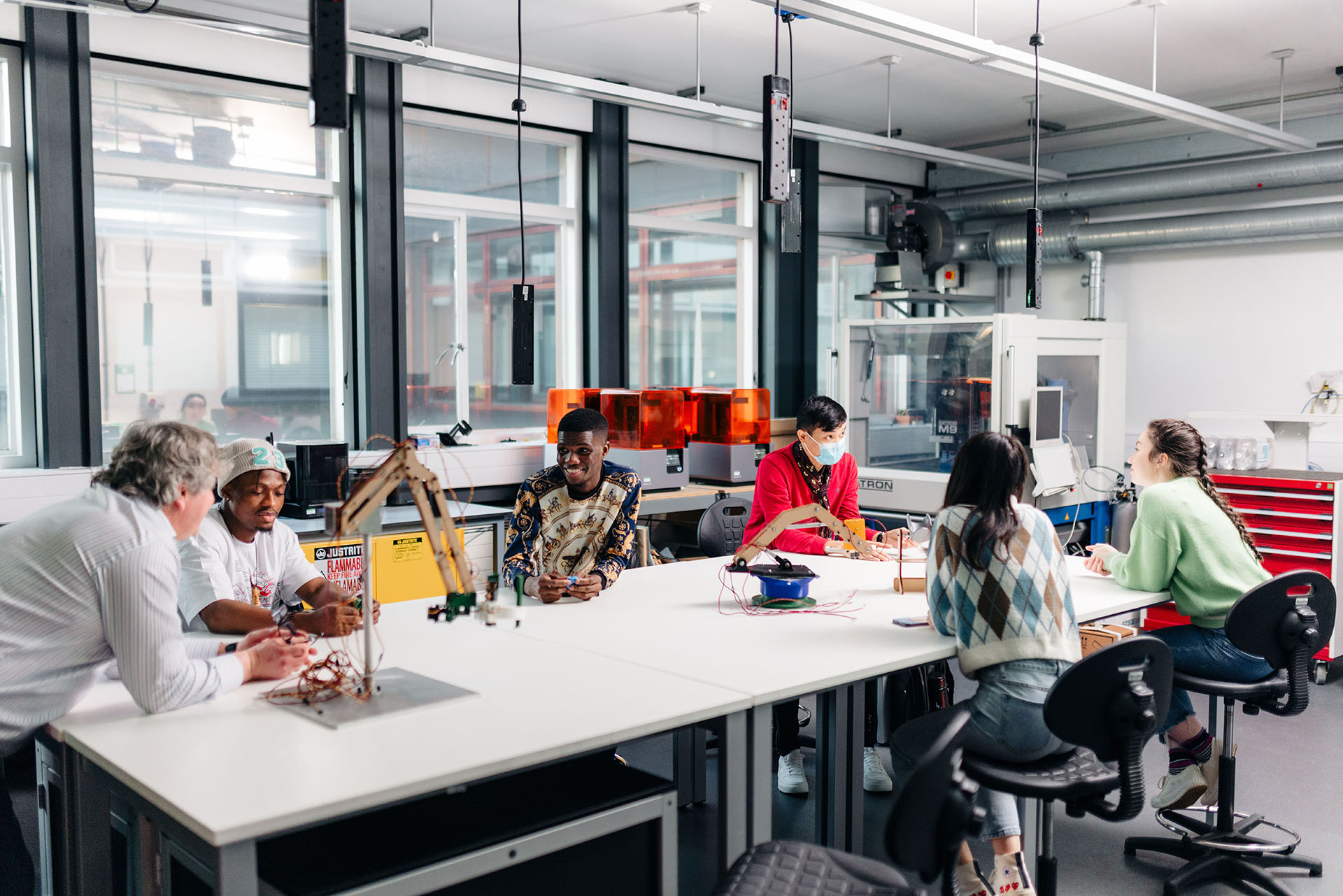 Students sit around a large white table in a brightly lit room surrounded by computer equipment 