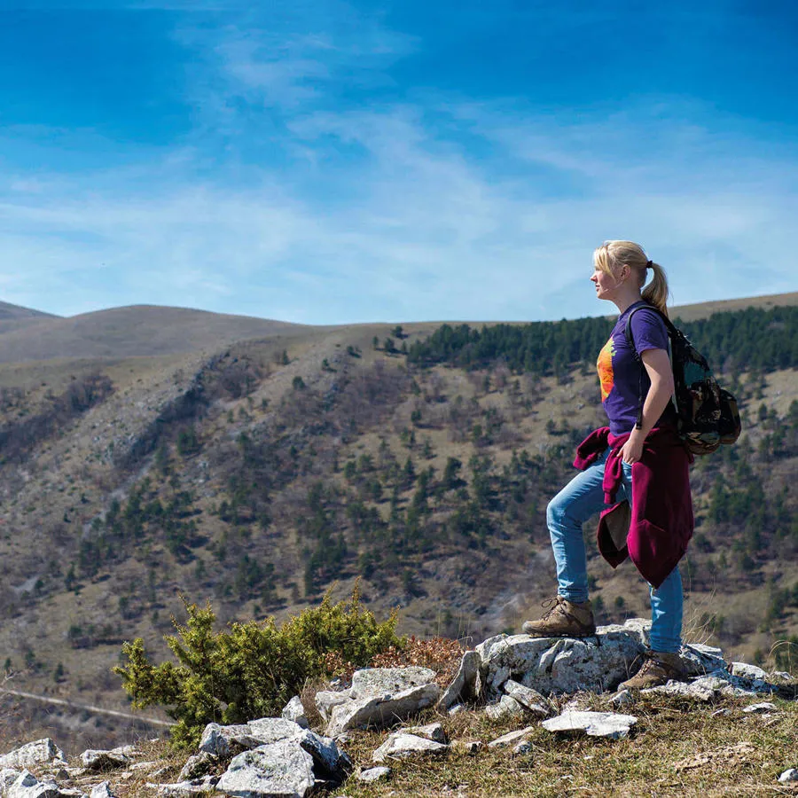 A young woman in casual clothing stands on the top of a mountain looking out over a cloudy blue sky