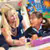 A female teacher helps a young girl with her work surrounded by bright, colourful pictures on the wall of a classroom.