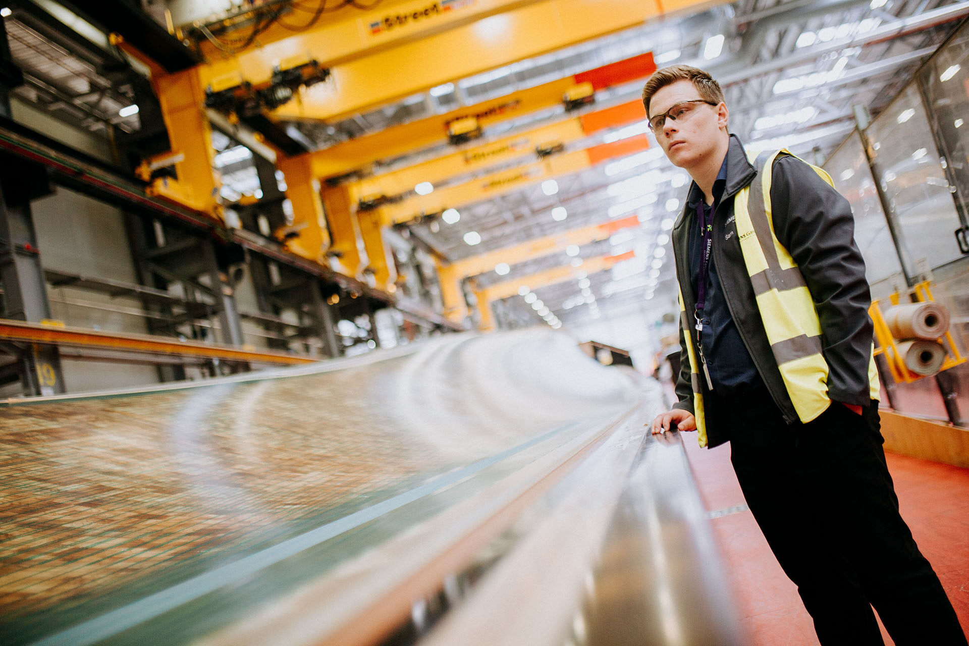 A young man in a high vis vest and safety goggles looks across an assembly line in a brightly lit factory