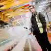 A young man in a high vis vest and safety goggles looks across an assembly line in a brightly lit factory